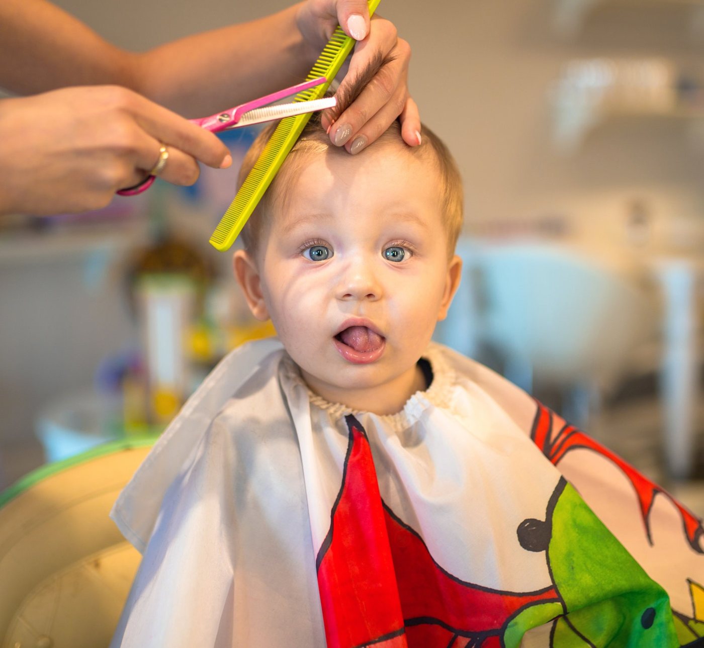 A toddler pulling a face in the chair, wearing a cape, while a stylist lifts a section of his hair with a comb and shears.