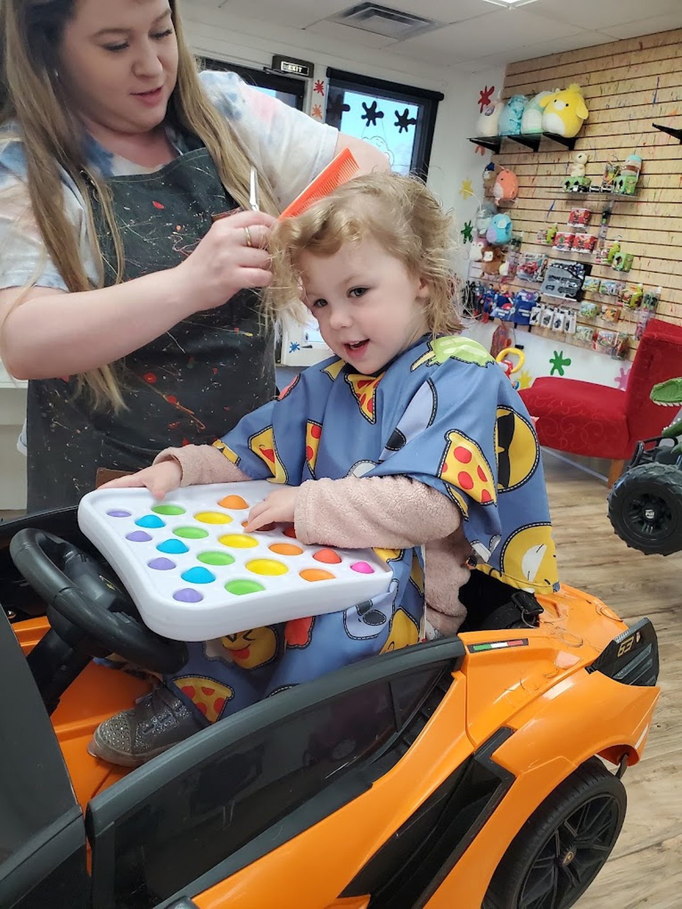 A toddler in an orange ride-on sports car holding a pop-it tray while a stylist trims her hair, the toy wall behind them.