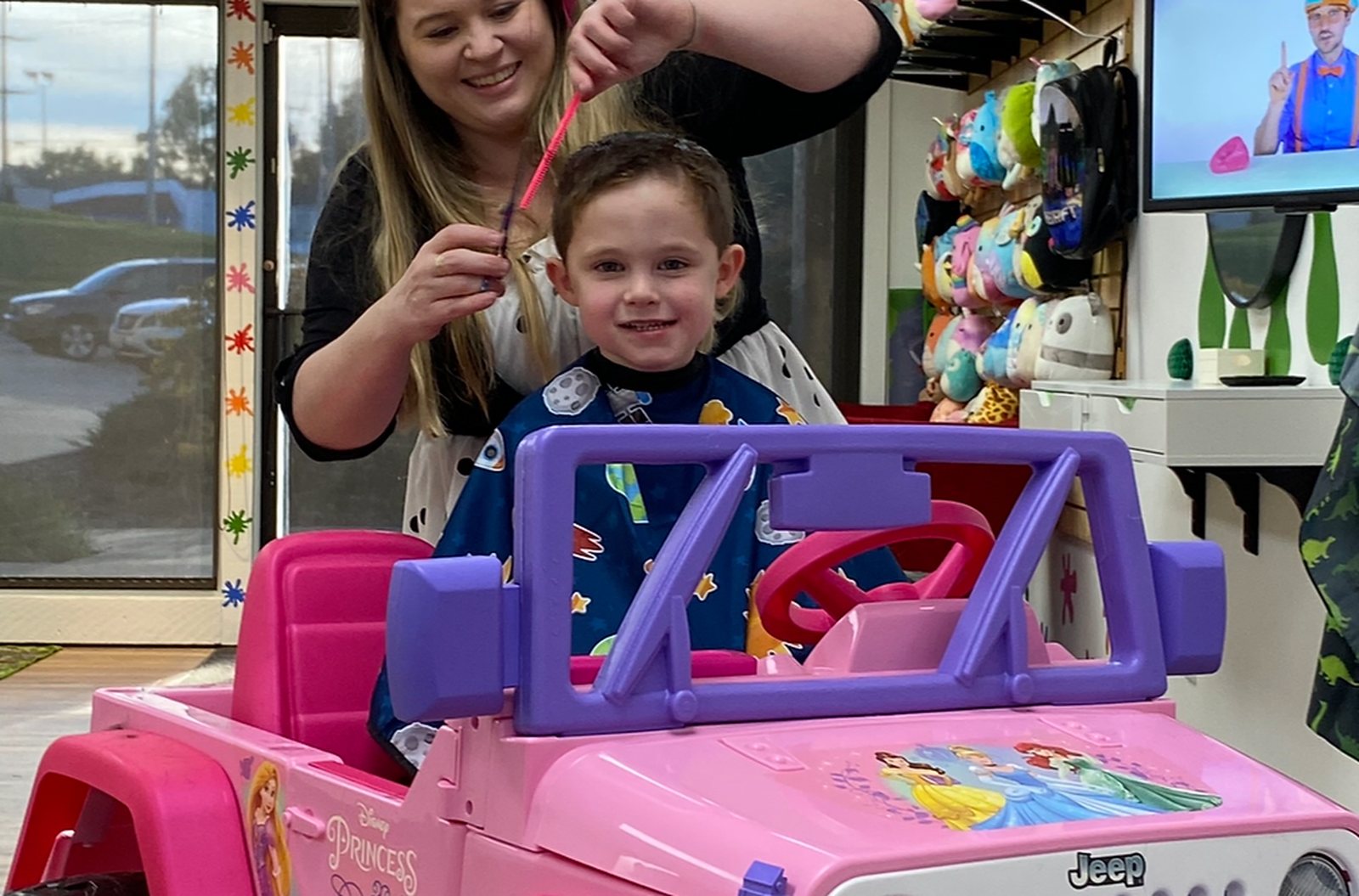 A child in a pink ride-on car by the front window while a stylist combs his hair, paint splat decals stuck to the storefront glass.